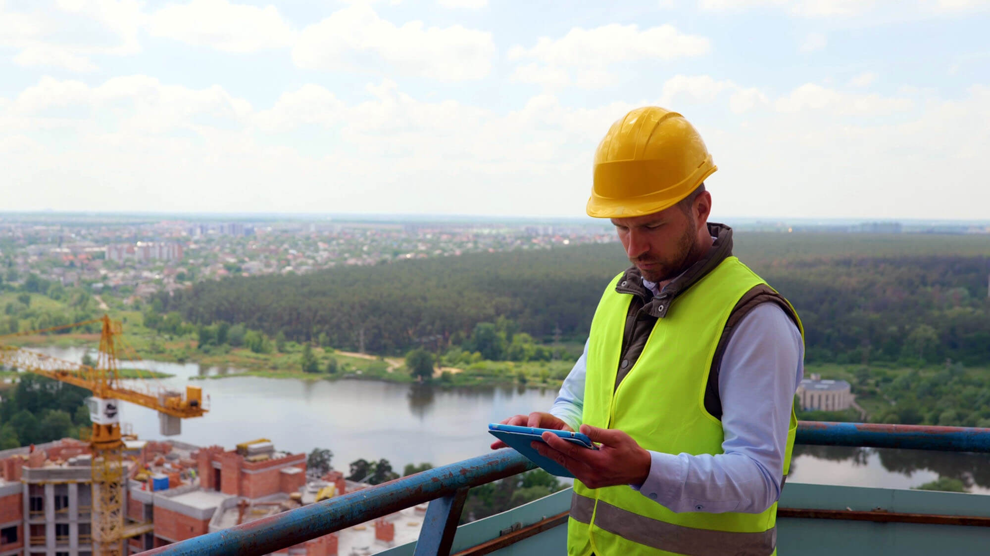 Focused Caucasian young man in uniform and helmet working on digital tablet while standing on construction site. Competent engineer checking working process. Building industry.